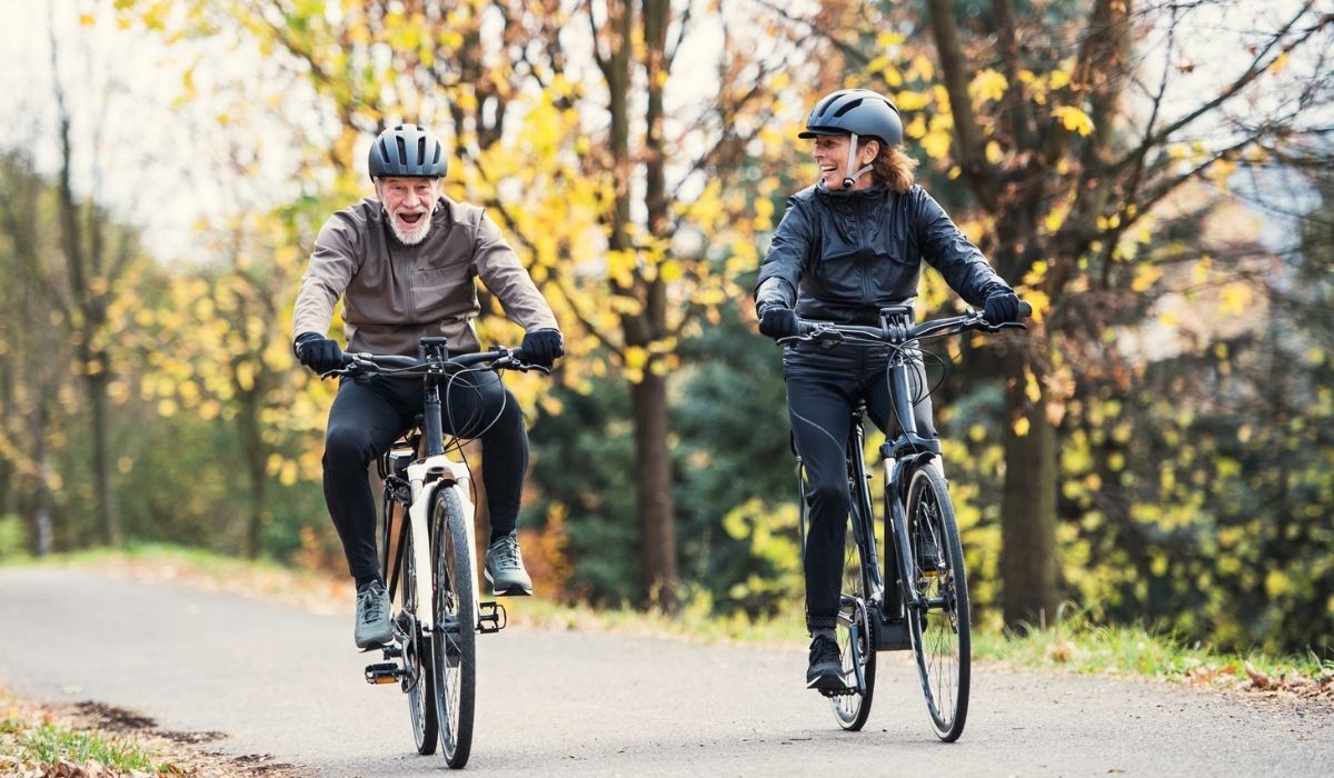 A senior couple with electrobikes cycling outdoors on a road in park in autumn