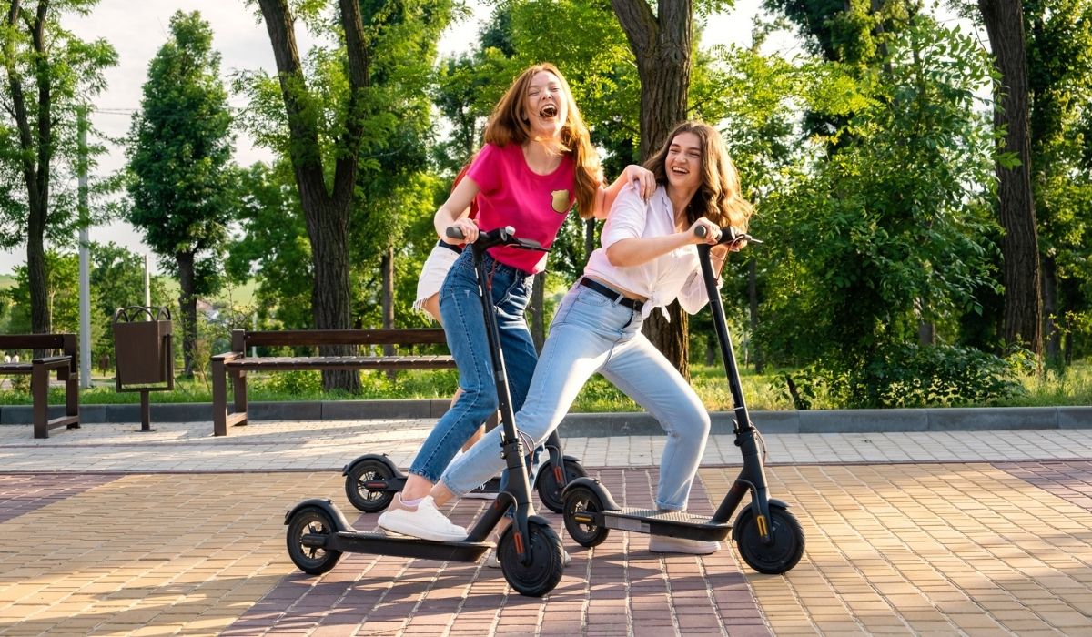 Three Young girl friends on vacation having fun driving electric scooter through the city park